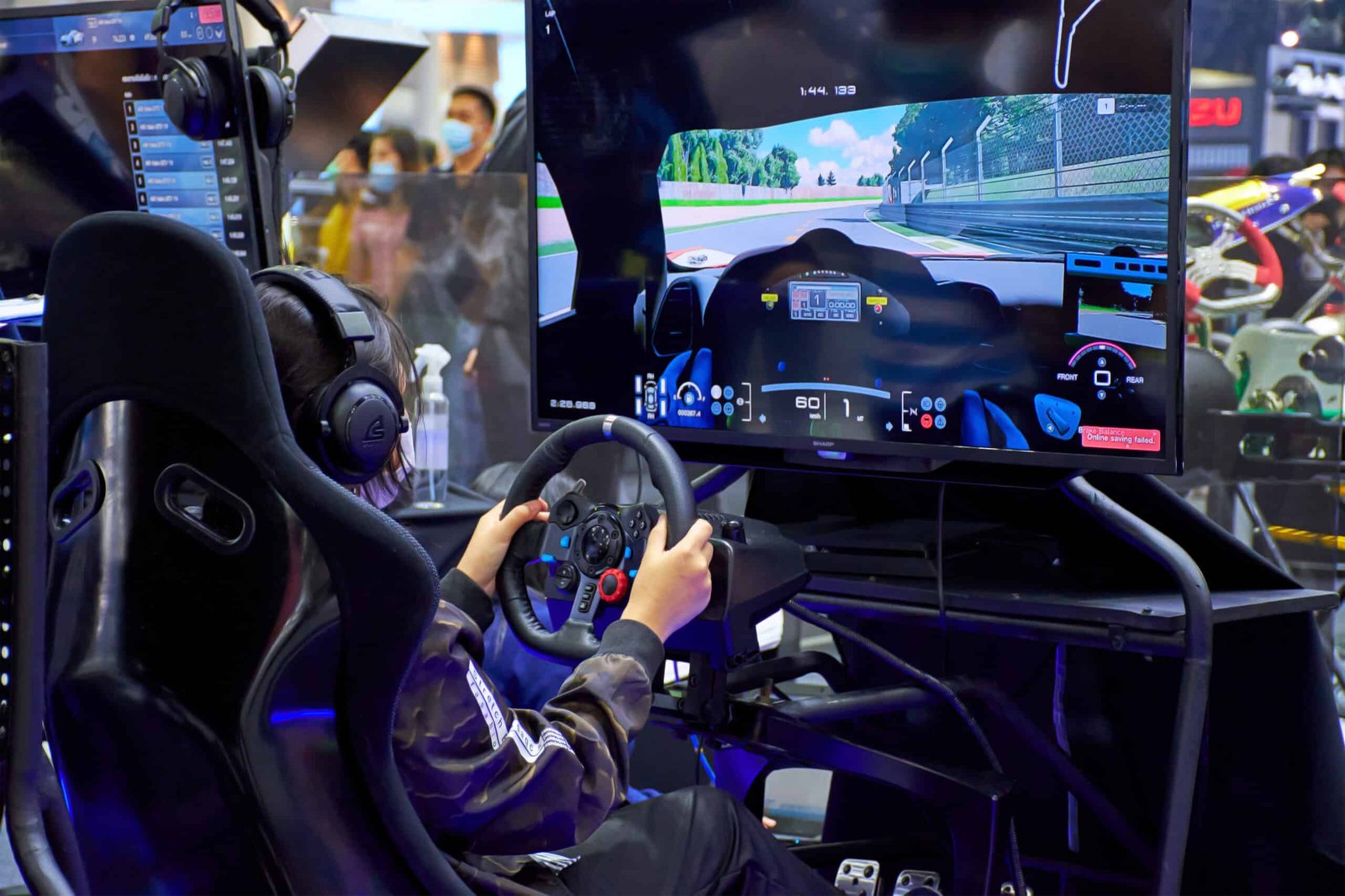 Jeune pilote équipé d'un casque audio au volant d'un simulateur, face à un écran affichant une vue cockpit sur circuit, dans un salon d'exposition
