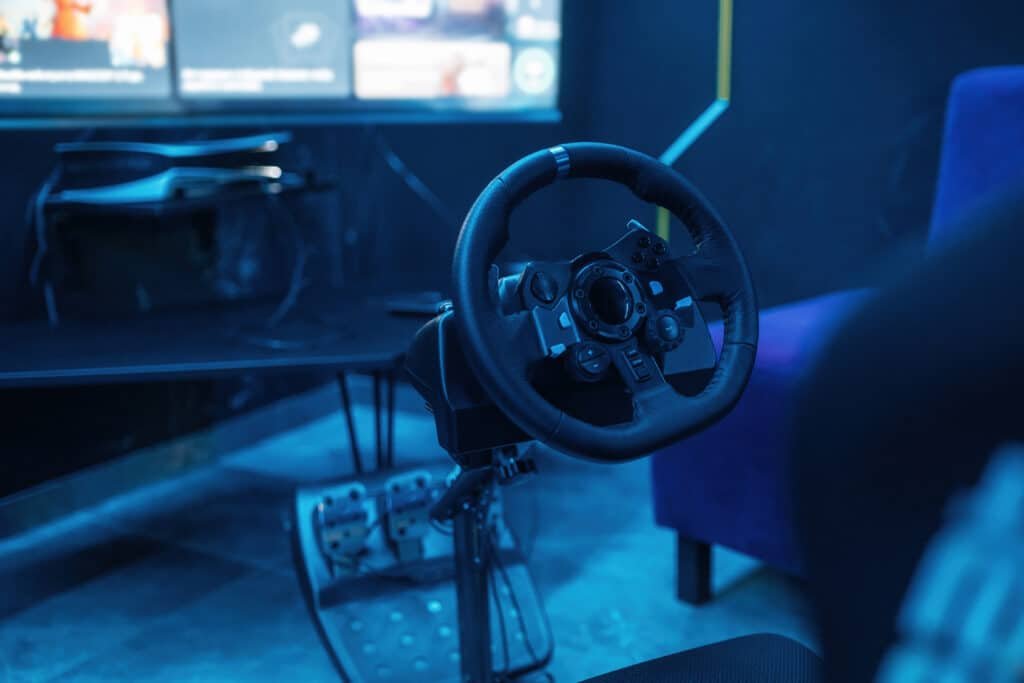 Volant simracing rond fixé au bord d'une table avec son pédalier au sol, pièce éclairée en bleu