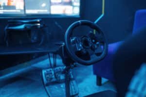 Volant simracing rond fixé au bord d'une table avec son pédalier au sol, pièce éclairée en bleu