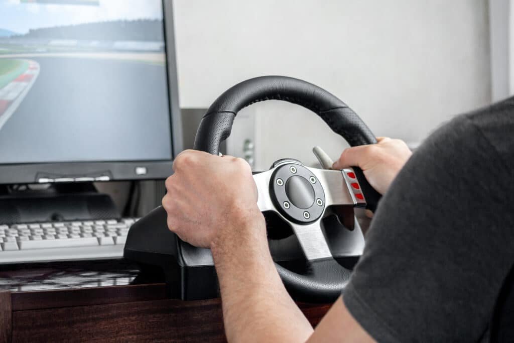 Mains d'un pilote posées sur un volant simracing rond à branches métal, fixé à un bureau devant un écran affichant un circuit
