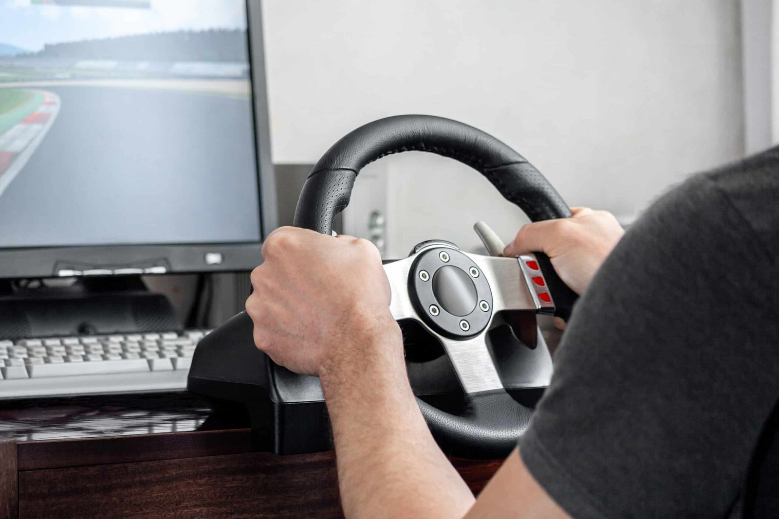 Mains d'un pilote posées sur un volant simracing rond à branches métal, fixé à un bureau devant un écran affichant un circuit